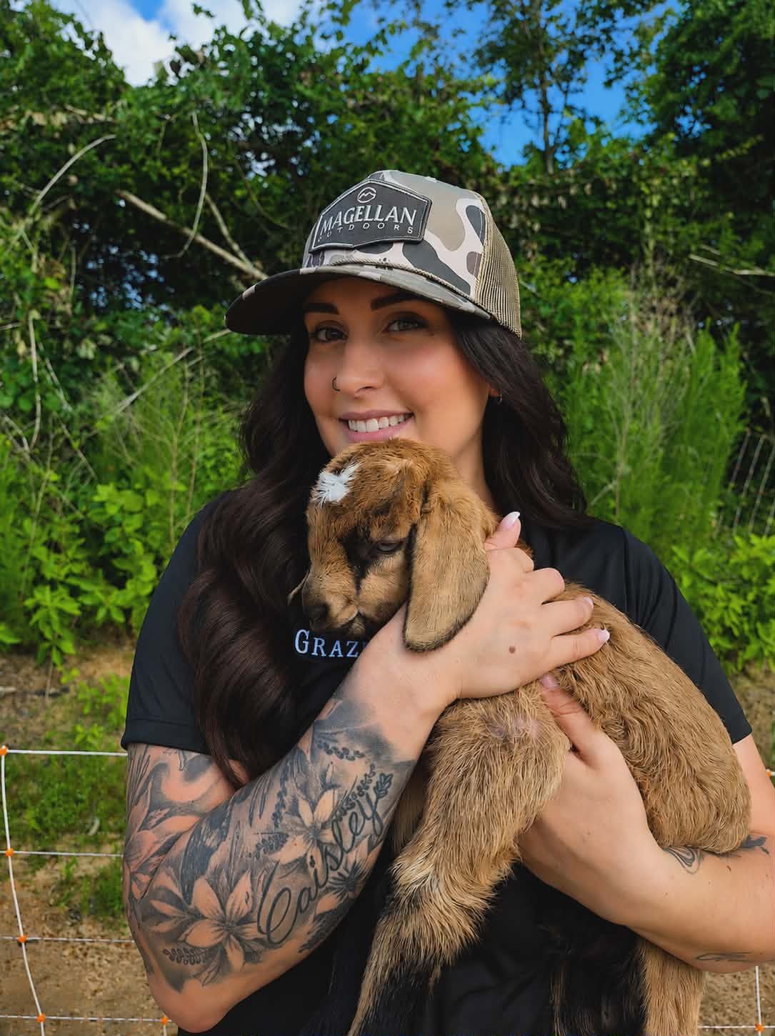 Woman with long dark hair and a tattooed arm smiles outdoors, holding a brown baby goat amid greenery near The Grazery restaurant at Two Step Farm.