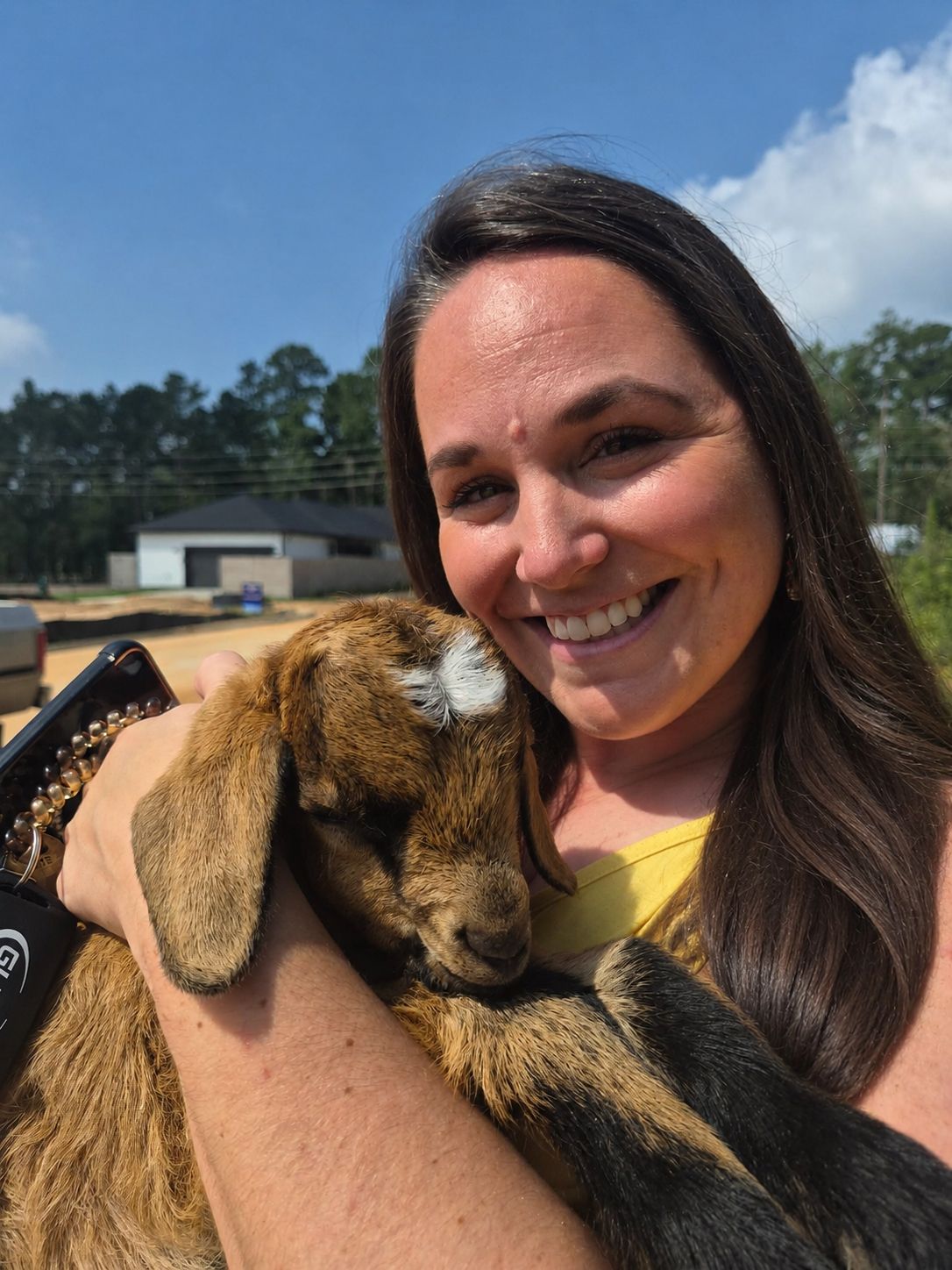 A woman smiles while holding a brown baby goat in her arms outdoors on a sunny day, enjoying the peaceful atmosphere near new homes at Two Step Farm, TX.