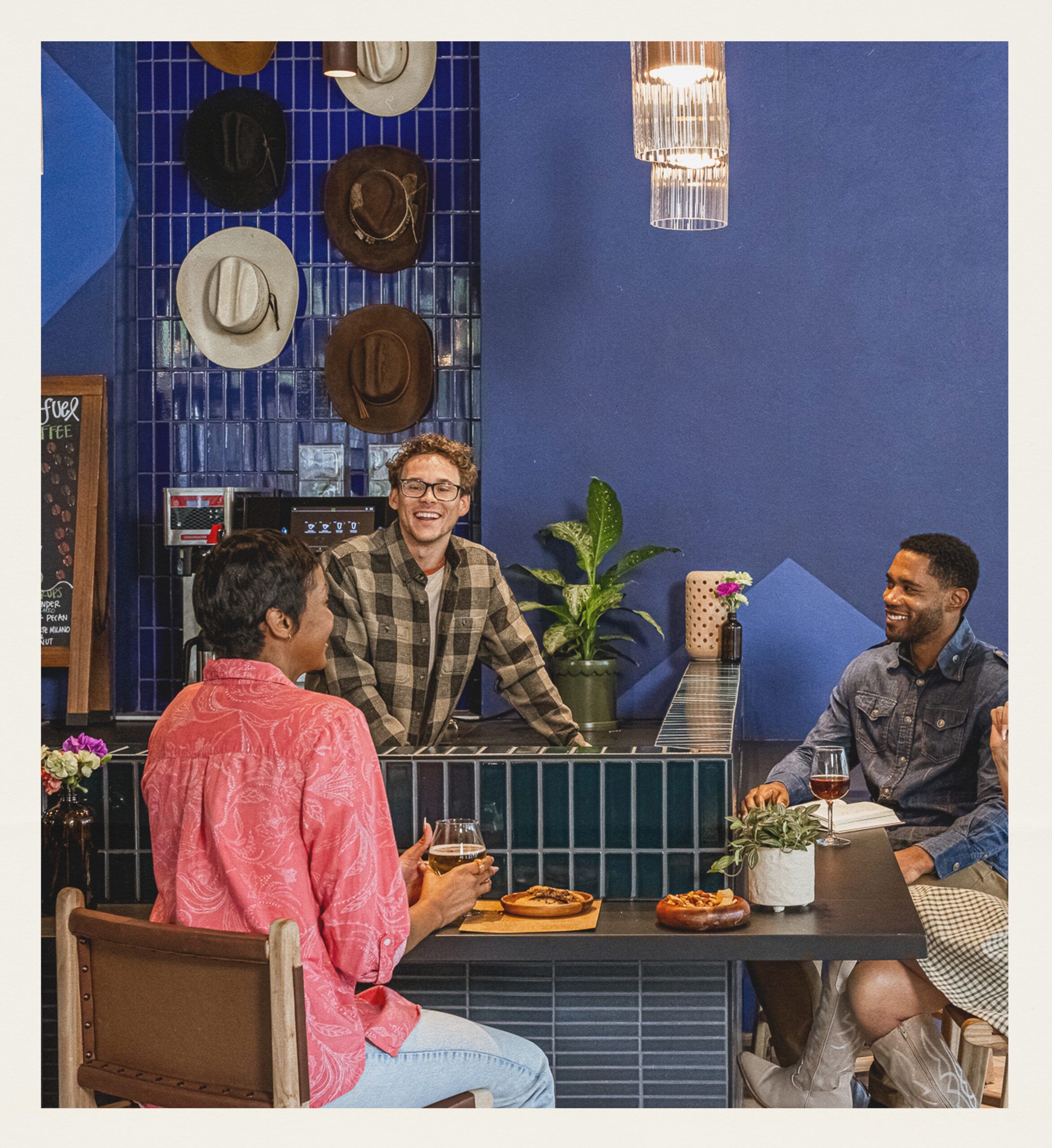 Three people sit at a table with food and drinks, talking and smiling in The Grazery restaurant at Two Step Farm—a blue-tiled café decorated with hats—perfect for those exploring new homes in Montgomery County, TX.