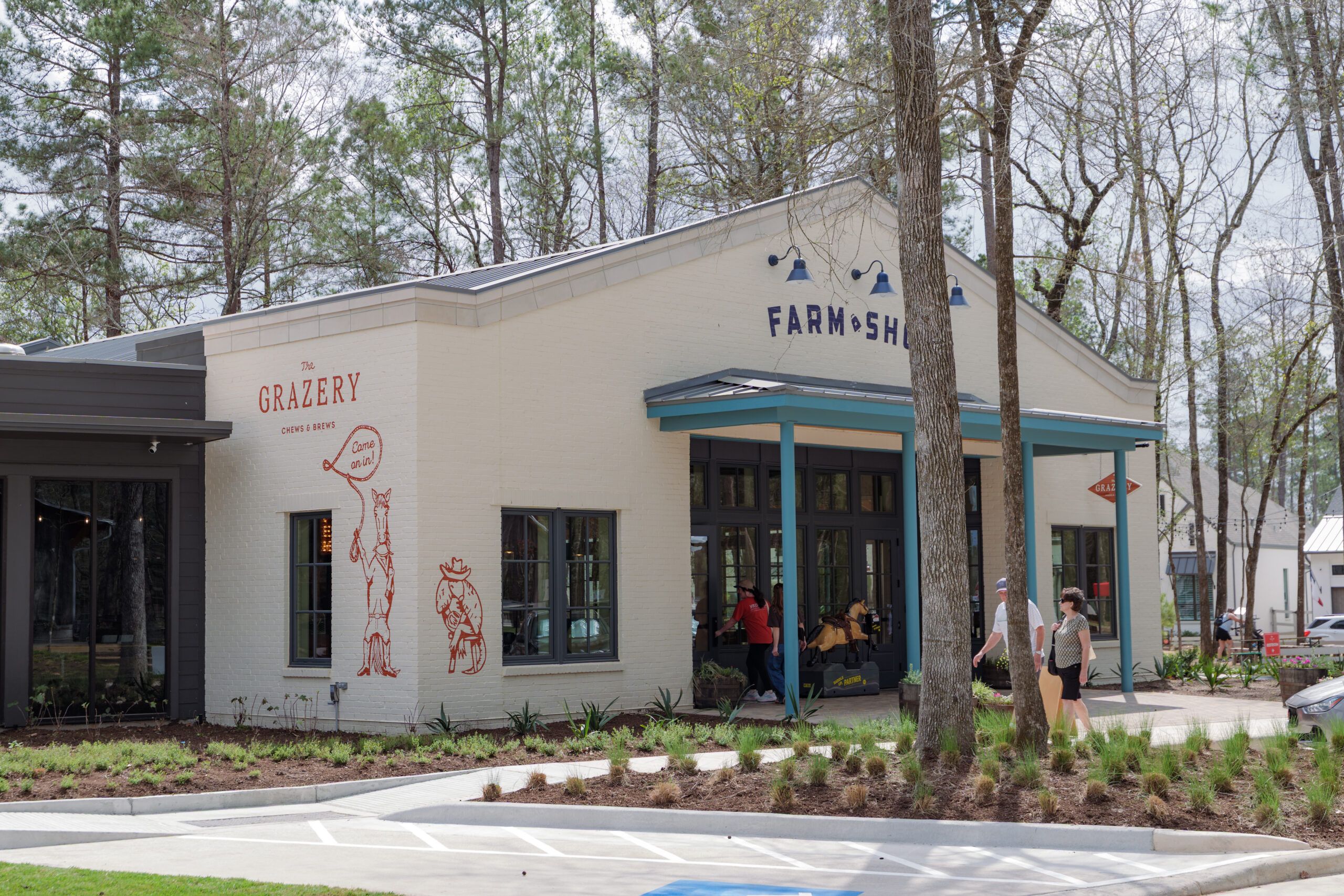 A white brick building labeled "Farmshop" with large windows, a side mural reading "The Grazery," and people entering and walking by—as seen near new homes in Montgomery County, TX, and The Grazery restaurant at Two Step Farm.
