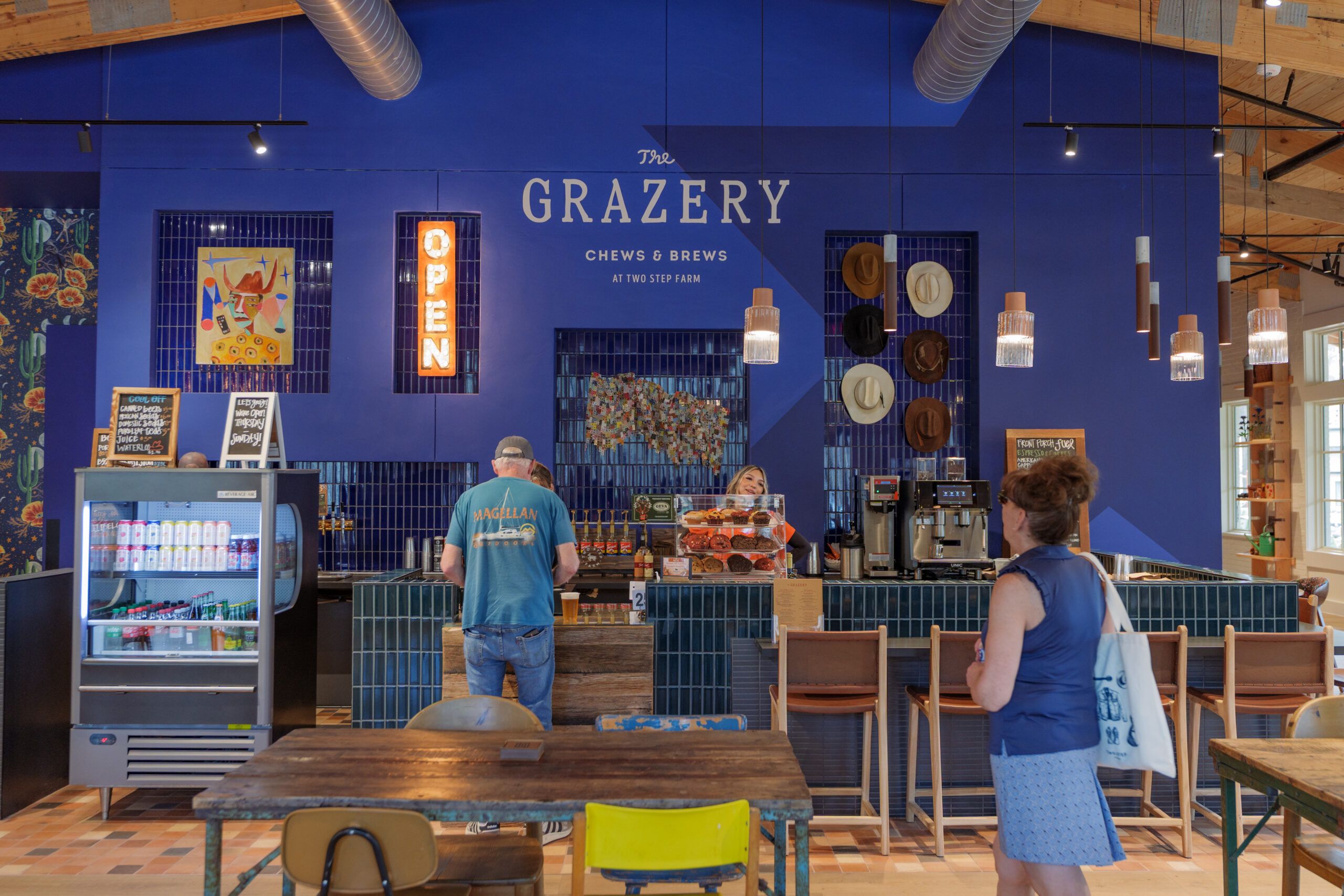 A man orders at the counter of The Grazery restaurant at Two Step Farm, with a display fridge, coffee machines, and a neon "OPEN" sign on a blue wall behind—capturing local TX charm. Other customers stand nearby.